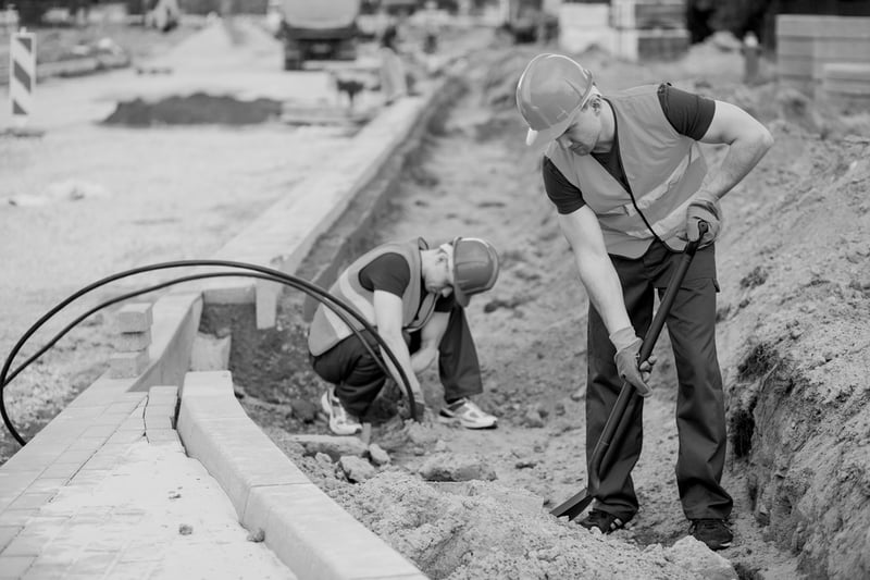 two construction workers with hardhats shoveling through dirt at roadside work site