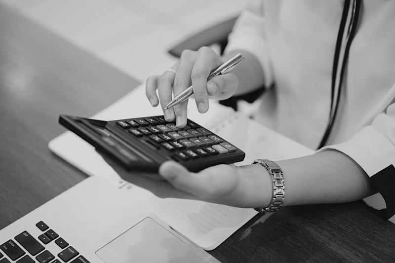 hands wearing a watch and holding calculator and pen at desk in front of notebook and laptop