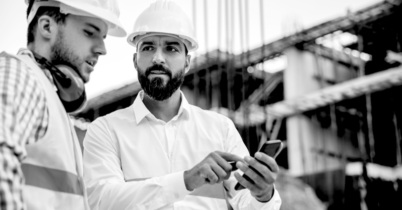 2 contractors with mobile phone in hand at a job site with a blurred image of construction happening in the background