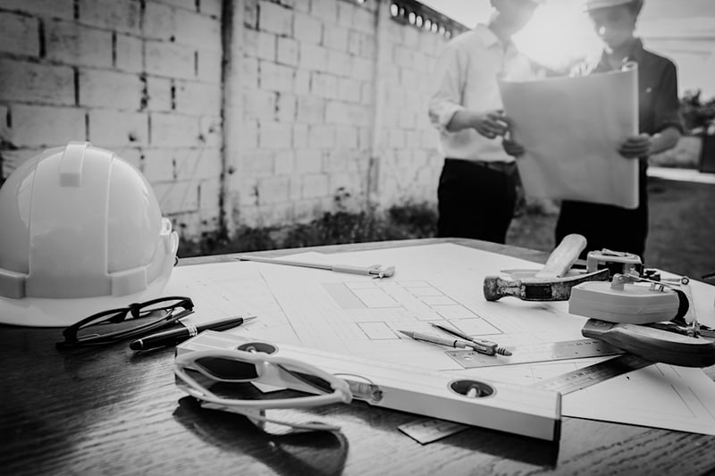 two construction workers reviewing job site plans in front of table with hardhat and drafting equipment