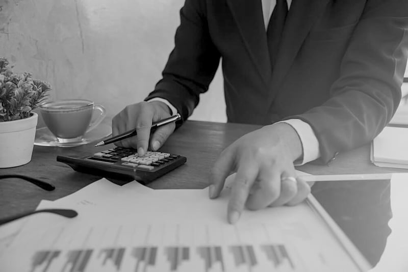 businessman in black suit at desk using calculator and pen while pointing at a printed data sheet