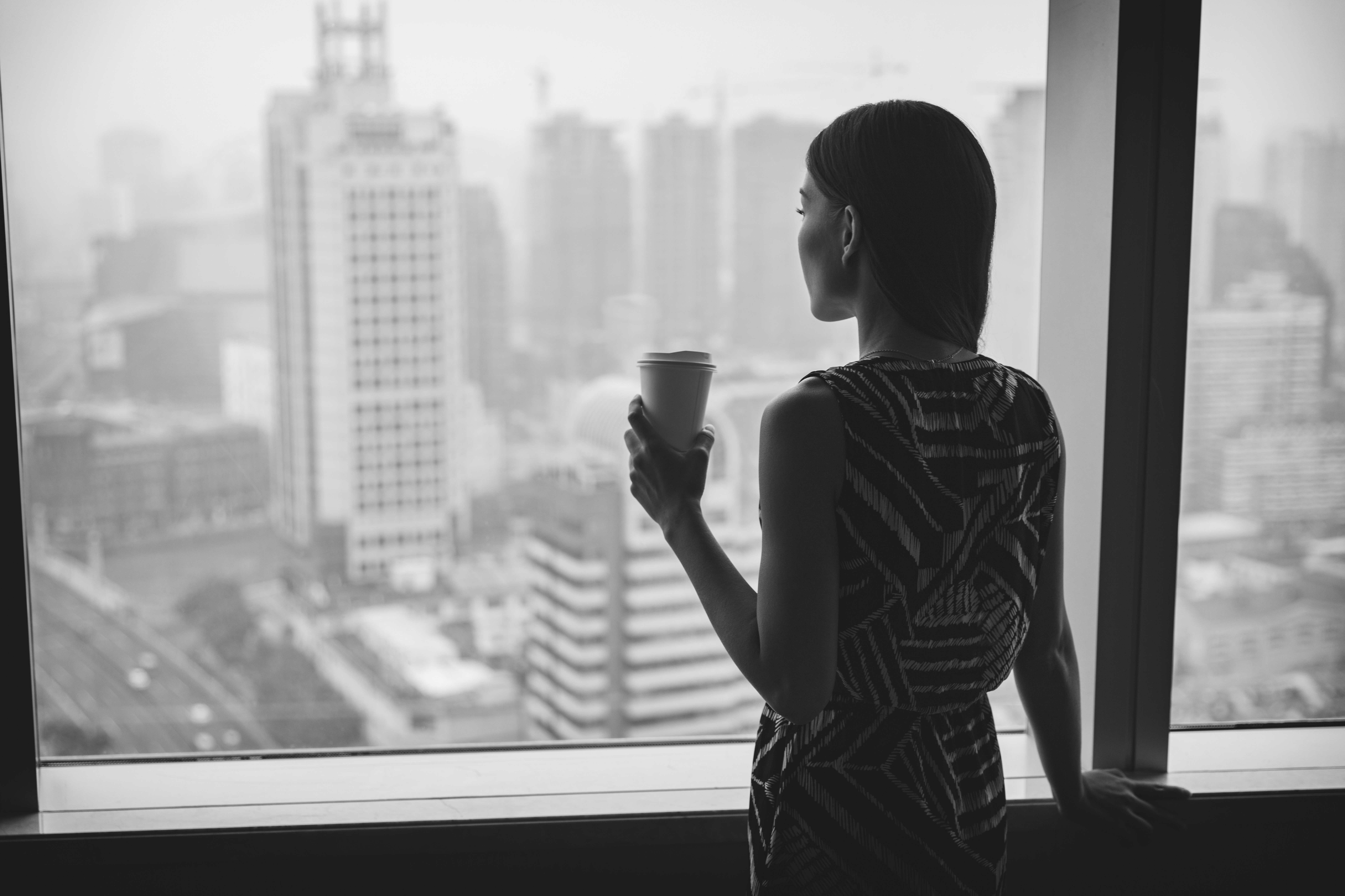 Businesswoman drinking coffee at work contemplative looking out the window of high rise skyscraper building.