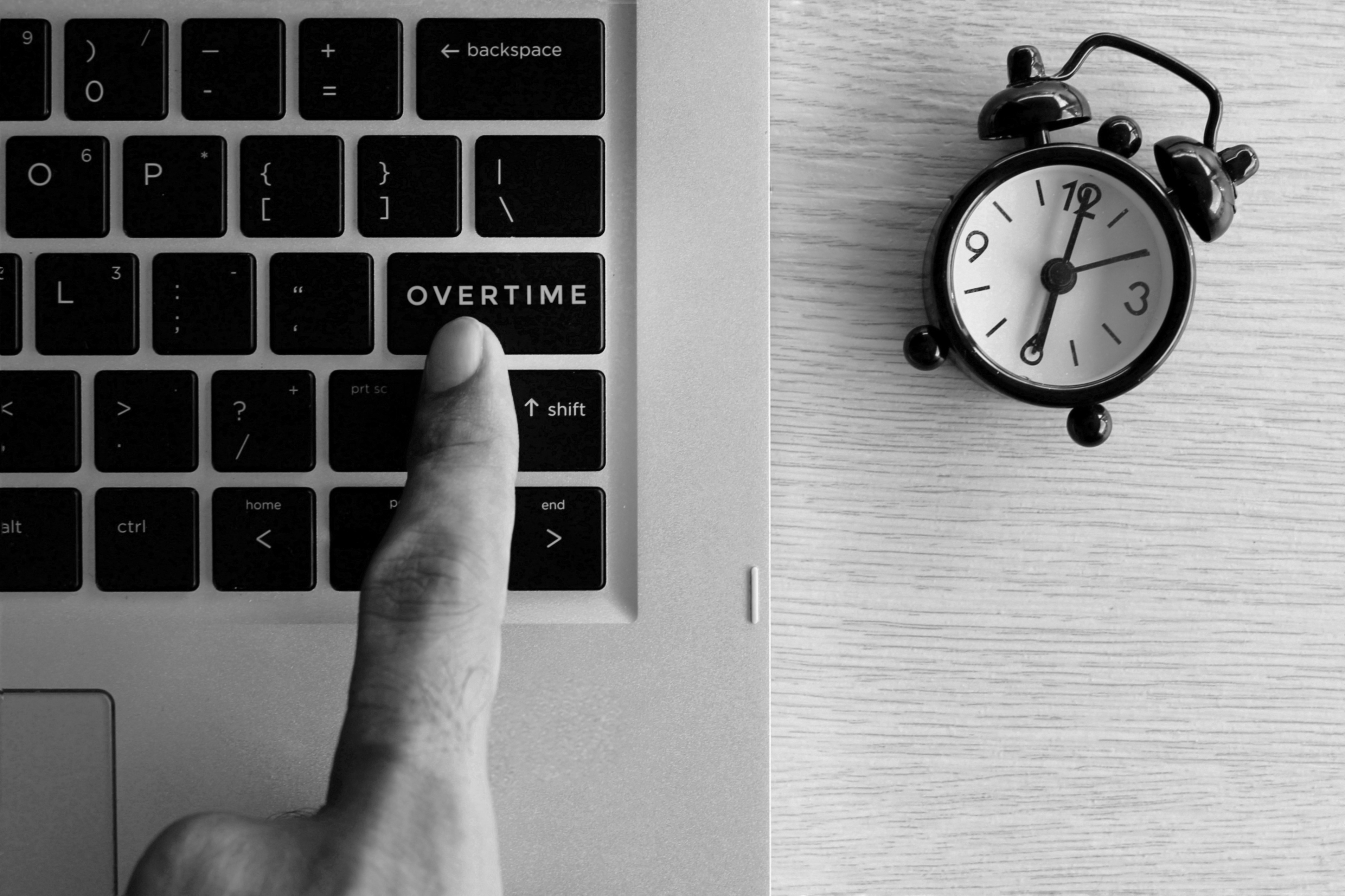 A finger pointing at a keyboard that says overtime with an alarm clock next to it.